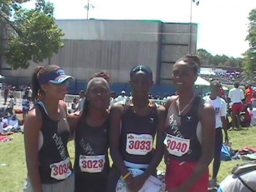 Group of female track athletes at a meet, wearing black uniforms with race numbers.