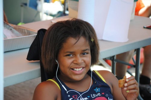 Young girl smiling at a track and field event, holding a snack and wearing headphones.