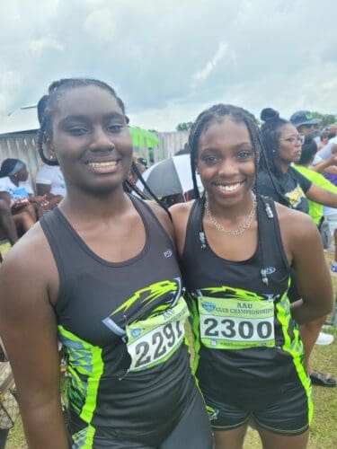 Two young female track athletes smiling at a meet, wearing black and neon green uniforms.