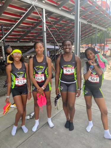 Girls track team at a meet, smiling and holding shoes.