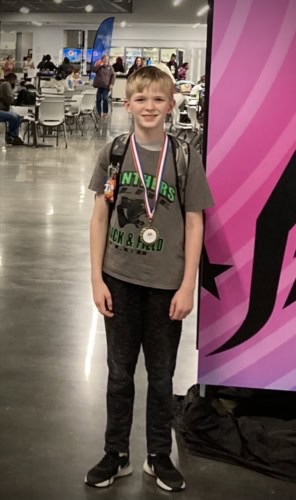 A young boy smiling with medals at a track and field event indoors.