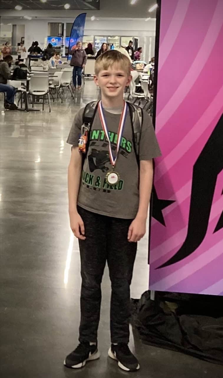 A young boy smiling with medals at a track and field event indoors.