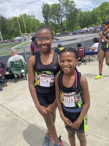 Young female athletes smiling at a track meet, wearing black and neon green uniforms.