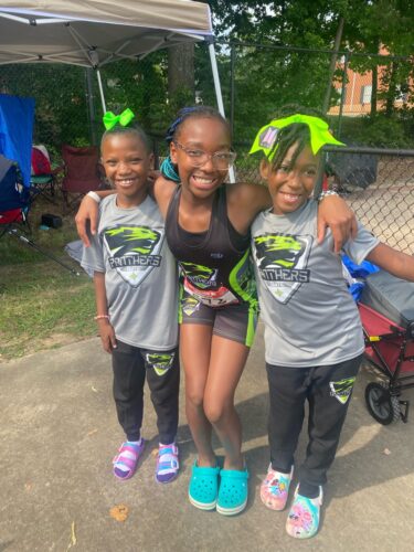 Young athletes smiling at a track event, wearing Panthers Elite gear and bright hair accessories.