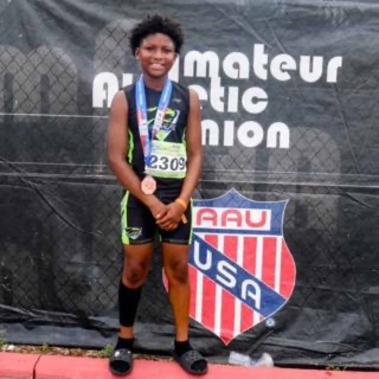 Young athlete with medals at a track event, standing in front of a Panthers Elite banner.