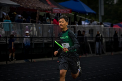 Young athlete running on track during a track meet, holding a relay baton.