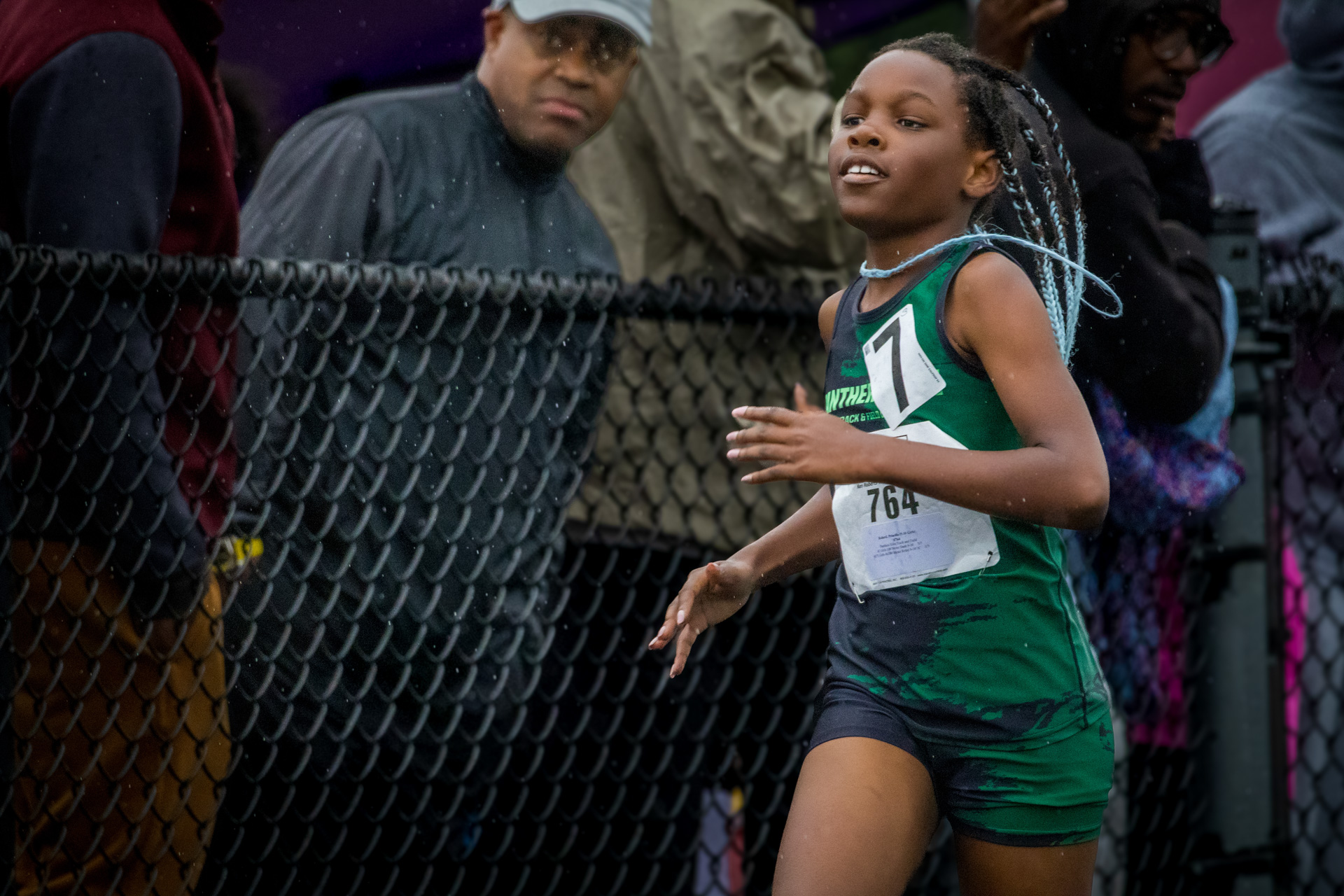 Athlete running during a track meet, with spectators watching behind a fence.