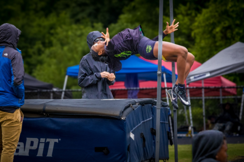 Athlete performing a high jump at a Panthers Elite Track & Field meet, with coach and spectators in.