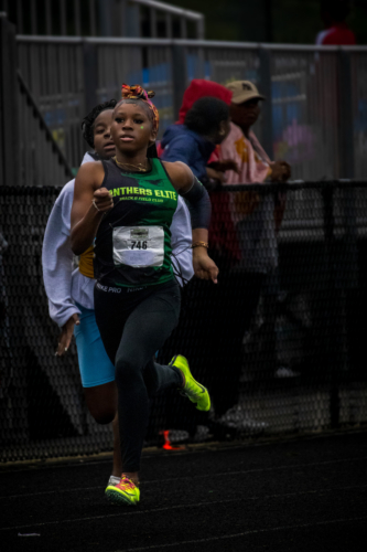 Young female athlete running on track during a meet, wearing Panthers Elite gear.