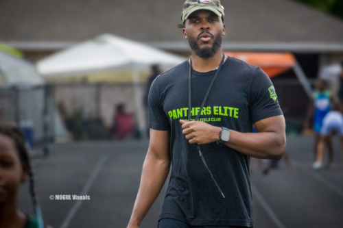 Panthers Elite track coach wearing a black T-shirt and cap at outdoor meet.