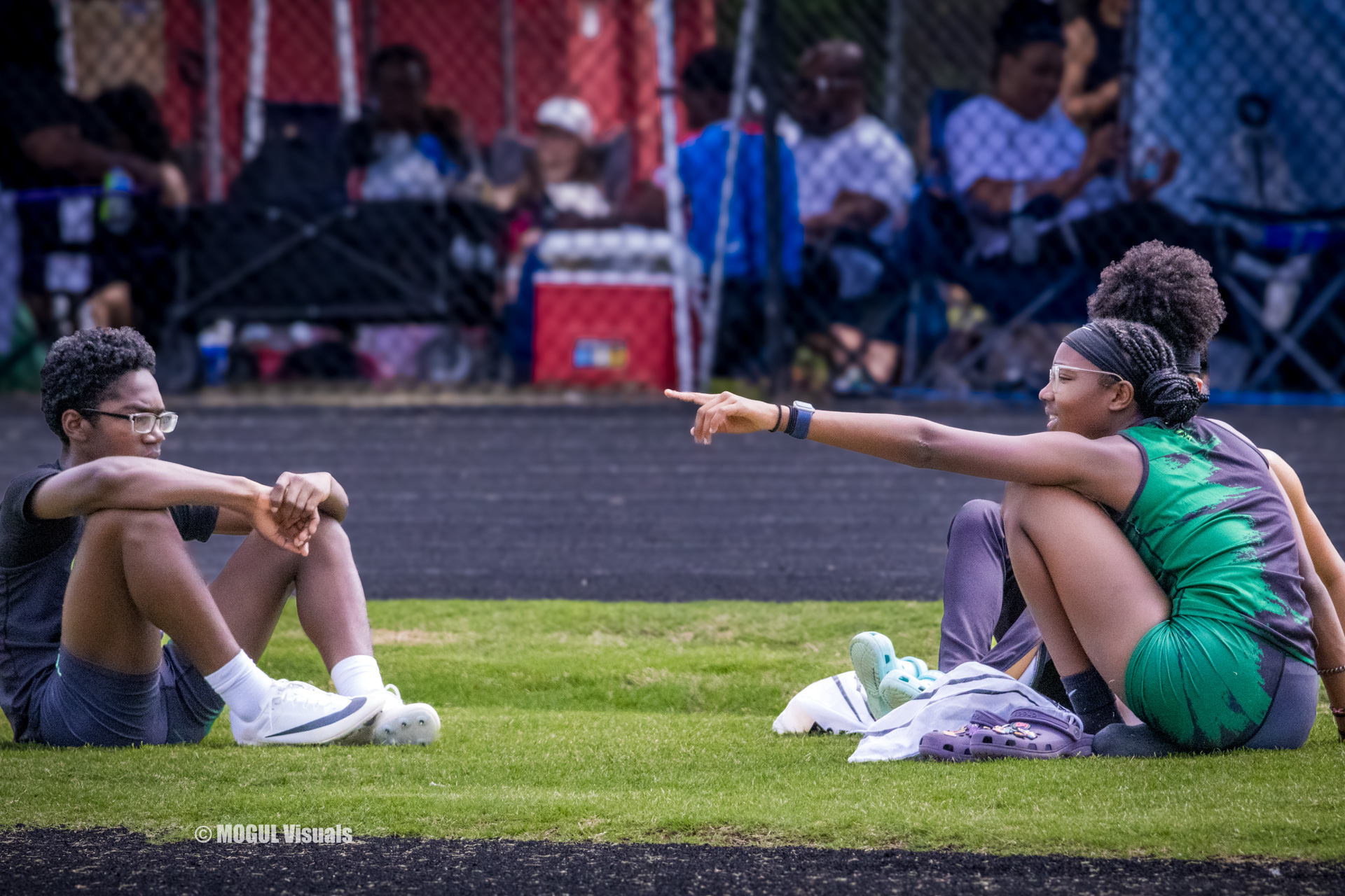Two young female athletes sitting on the track, stretching and talking during a meet.