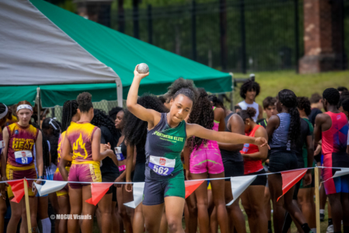 Young female athlete in green uniform preparing to throw a shot put at a track meet.