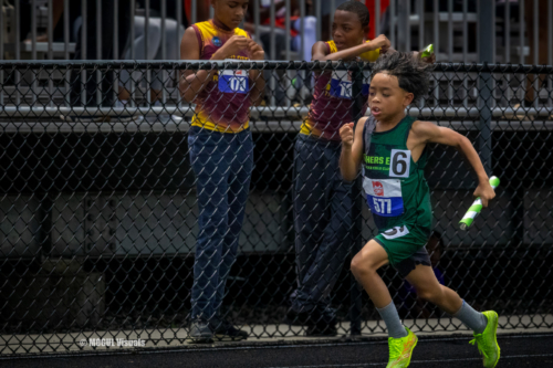 Young boy running in a track race, wearing green and black uniform with a baton.