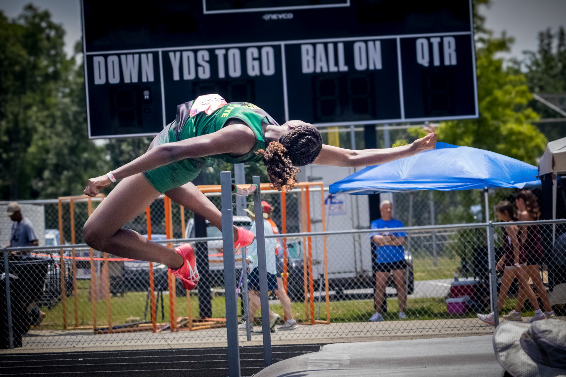 Female athlete in green uniform jumping over hurdle at track meet.