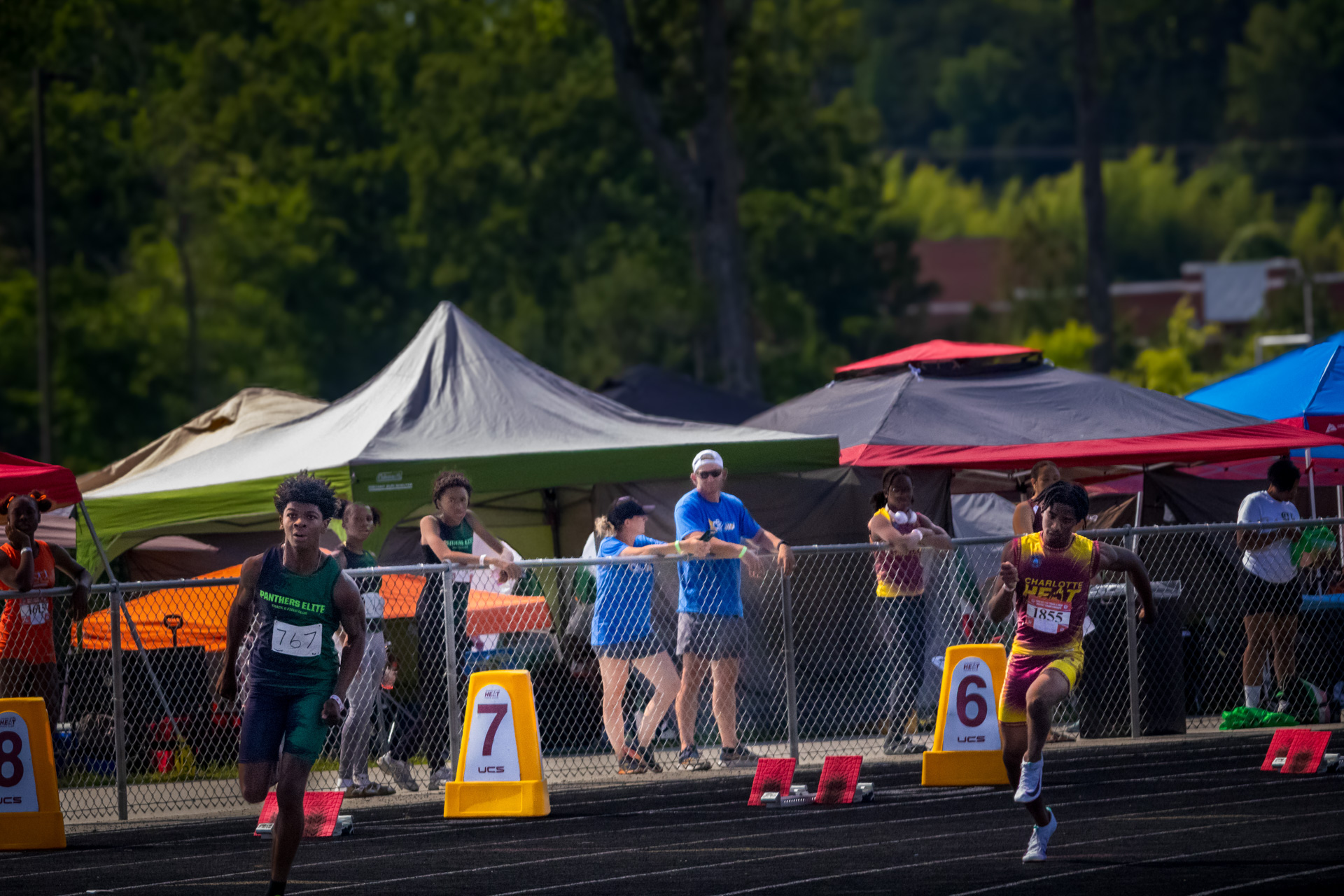 Track athletes competing at District Qualifier 2026 with tents and spectators in background.