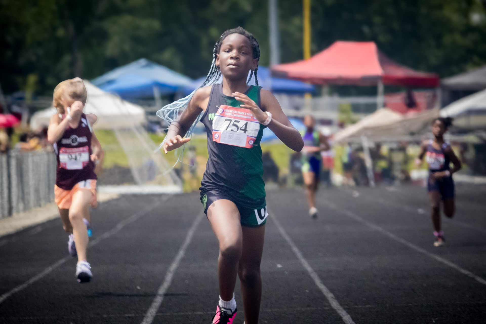 Young female sprinter running on track during district meet with other athletes in background.
